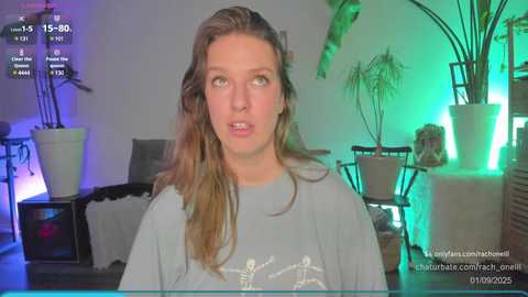 Media: Video of a young woman with long, wavy brown hair, wearing a gray T-shirt, standing indoors with green and purple ambient lighting, surrounded by potted plants and furniture.