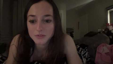 Media: Video of a young woman with long brown hair, wearing a black top, in a dimly lit bedroom with a bed covered in a zebra-patterned blanket.