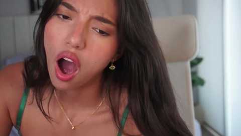 Video of an Asian woman with long black hair, wearing a green bra, open-mouthed, in a white chair, indoors.