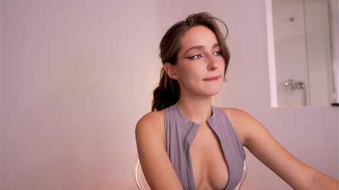 Media: Video of a Caucasian woman with fair skin, brown hair, and green eyes, wearing a low-cut, sleeveless gray dress, smiling softly. Background features a white wall and a partially visible bathroom door.