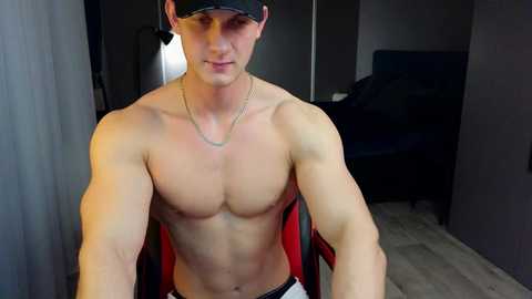 Media: Video of a shirtless, muscular young man with fair skin, wearing a black cap, necklace, and white pants, seated in a modern room with a bed in the background.