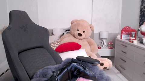 Media: A video of a plush teddy bear sitting on a gray gaming chair in a minimalist, white-walled room with a beige couch, red pillow, and Santa Claus decorations on a white dresser.