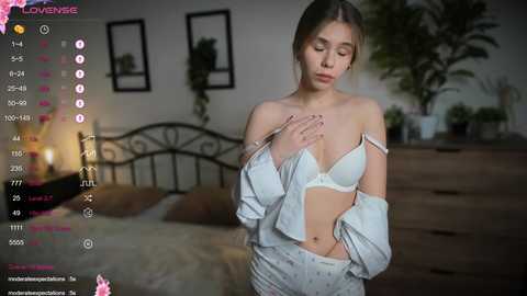 Media: A video of a young woman in white lingerie, partially unbuttoned, standing in a dimly lit bedroom with a bed and potted plants in the background.