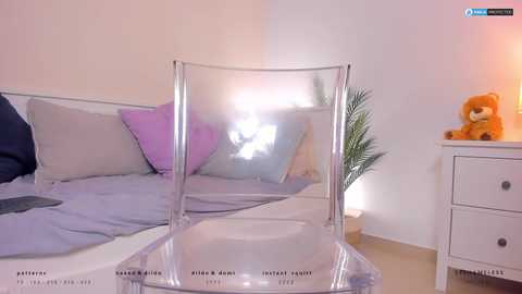 Media: Video of a minimalist bedroom with a clear plastic chair, a white dresser, and a teddy bear, featuring soft pastel bedding and a bright light source.