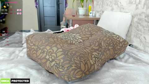 A video shows a woman in bed with a brown, floral-patterned duvet, a white pillow, and a gray blanket. The room features a gray and white patterned wallpaper, a wooden nightstand with a lamp, and a potted plant.