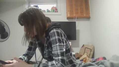 Media: A video of a young person with shoulder-length brown hair, wearing a black and white plaid shirt, seated at a desk, looking down. The room has a white wall, a window with a small plant, and a flat-screen TV.