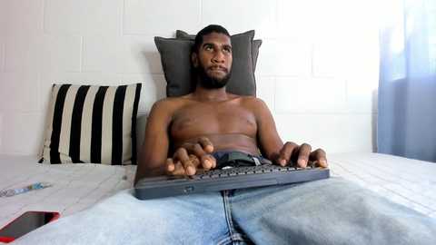 Media: Video of a shirtless Black man with a trimmed beard, seated on a bed, typing on a laptop, with a red phone and striped pillow beside him.