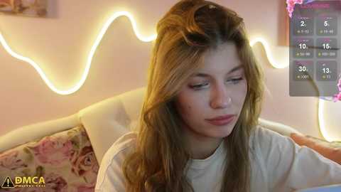 Media: Video of a young woman with long brown hair, fair skin, and a contemplative expression, sitting on a pastel floral couch. Soft, warm ambient lighting creates a cozy, inviting atmosphere.