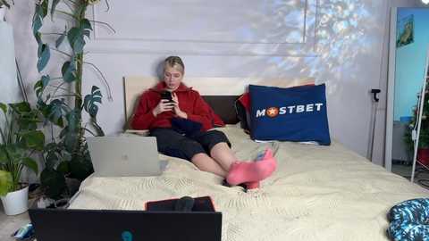 Media: Video of a blonde woman in a red hoodie, black shorts, and pink socks, sitting on a bed with a beige blanket. Laptop, blue pillow, and green plants in background.