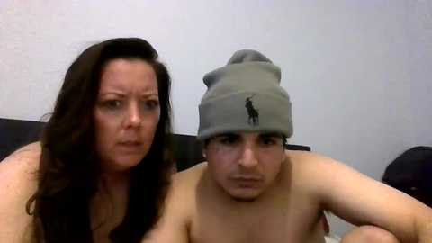 Media: Video of a fair-skinned woman with long dark hair and a man with a grey beanie, both topless, seated on a bed with a white wall background.