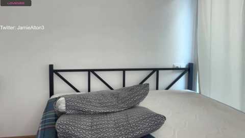 Media: Video of a minimalist bedroom with a black metal headboard, white walls, a white bed with grey patterned pillows, and a sheer curtain on the right.