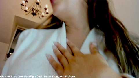 Media: A video capturing a close-up of a woman with fair skin, long brown hair, and a white shirt. Her hand gently touches her chest. The background features a chandelier and a beige wall.