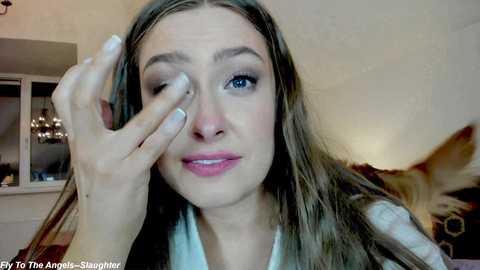 Media: Video of a young woman with long brown hair, blue eyes, and fair skin, winking and applying eyeliner with her left hand. Background shows a cozy living room with a beige wall, a window, and a patterned cushion.