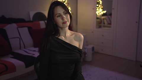 Media: Video of a fair-skinned woman with long black hair, wearing a black off-shoulder dress, standing in a dimly lit, cozy living room with a white cabinet, red couch, and fairy lights.