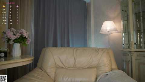 Media: Video of a dimly lit, cozy living room with beige leather couch, white vase of pink flowers on a side table, and a lit table lamp beside a glass cabinet.