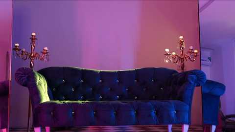 Media: Video of a dark green, tufted velvet sofa with a green velvet armchair, both illuminated by gold chandeliers with lit candles on either side, set against a purple-pink wall in a dimly lit room.