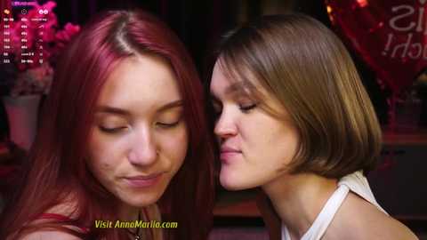 Media: Video of two young women with fair skin and long, straight hair, one with red, the other with brown. They're close, eyes closed, smiling softly. Background is dimly lit, with blurred festive decor.