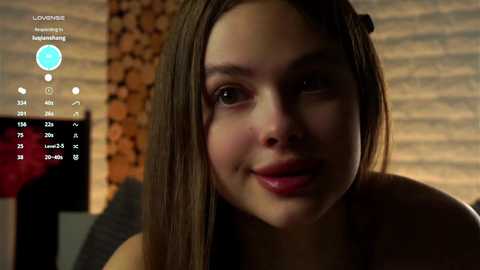 Media: Video of a smiling young woman with straight, brown hair, wearing a black top, in a dimly-lit room with brick wall and a flat-screen TV.