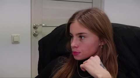 Media: Video of a young Caucasian woman with long, light brown hair, sitting pensively in a black leather chair, wearing a black top, with a visible tattoo on her wrist. The background features a white door and light switch.