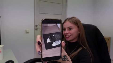 Media: Video of a smiling young woman with long brown hair, holding a smartphone showing a black-and-white photo of a person in a white shirt. She's seated in a black office chair, wearing a black turtleneck and brown apron, with a white wall and door behind her.