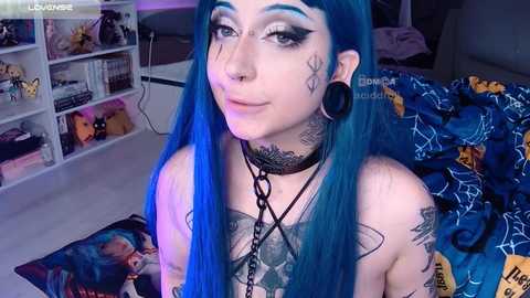 Media: Video of a young woman with vibrant blue hair, wearing black leather choker, heavy makeup, tattoos, and black ear gauges, in a messy bedroom with colorful bedding and shelves.