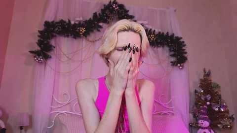 Media: Video of a slender, pale-skinned, blonde-haired person with a sleeveless pink top, covering their eyes, against a festive, decorated background with a Christmas tree and garlands.