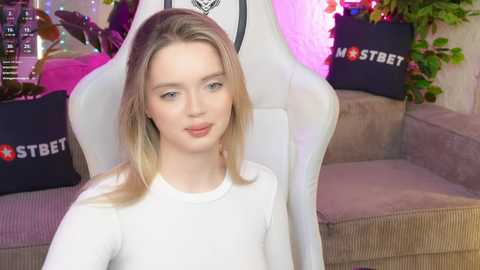 Video of a young, fair-skinned woman with shoulder-length blonde hair, wearing a white long-sleeved top, seated on a plush, white gaming chair with a black \"Strebet\" logo.