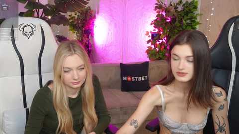 Video of two young women sitting on gaming chairs; one blonde in a green top, the other brunette in a lace bralette, both with tattoos. Background features a plant and \"Master Street\" pillow.