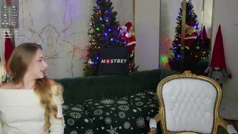 Media: Video of a blonde woman in a white sweater facing a green sofa with a white chair, Christmas tree with colorful lights, and \"Mastbet\" sign in the background.