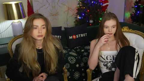 Media: Video of two young women with long hair, one wearing black and the other a white tank top, sitting on a couch with a festive background.