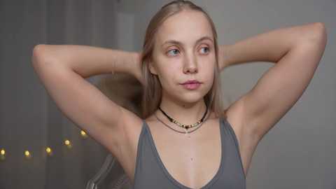 Media: Video of a young Caucasian woman with fair skin and light blonde hair, wearing a gray tank top, standing in a dimly lit room with a string of warm lights in the background.