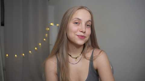 Media: Video of a young, fair-skinned woman with long, straight, light brown hair, wearing a gray tank top and multiple layered necklaces. She has a subtle smile and is indoors against a light-colored wall with softly glowing fairy lights.