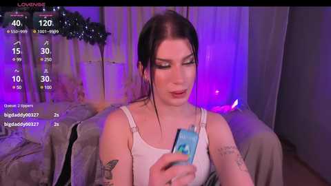 Media: A video of a young woman with pale skin, dark hair, and tattoos, wearing a white tank top, sitting in a dimly lit room with purple lighting, holding a smartphone.