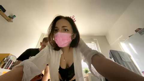 Media: A video shows a young woman with a pink mask, short brown hair, and a white shirt, taking a selfie in a cluttered bedroom with a window and a white door.