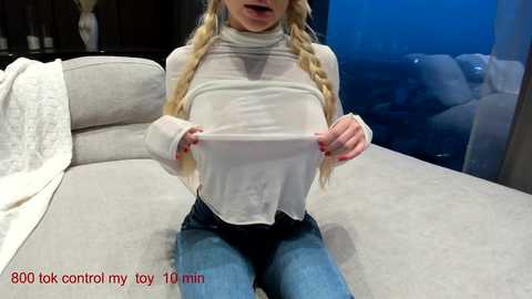 Media: Video of a slender, blonde woman with braided hair, wearing a sheer white top, lifting her shirt to reveal her small breasts. She sits on a grey sofa in a modern, dimly lit room.