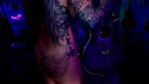 Media: A video of a tattooed person with long hair, holding a guitar, in a dimly lit, blue-lit room. The individual's tattoos cover their arms and chest, with the guitar displaying a skull emblem.