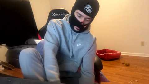 Media: Video of a person wearing a black ski mask and grey sweatpants, kneeling on a wooden floor in a dimly lit room with a red dog bed and a TV in the background.