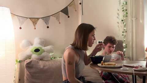 Video of a cozy living room scene: a young woman with shoulder-length brown hair, wearing a sleeveless gray top, sits on a beige sofa. A young boy, with curly brown hair, sits at a wooden table, eating. The room is decorated with festive banners, a stuffed toy, and fairy lights.