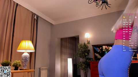 Video of a modern living room with beige walls, a lamp on a wooden table, potted plants, and a woman in a blue dress with white hair in the background.