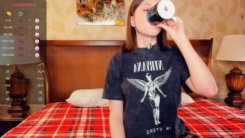 Media: Video of a woman drinking from a coffee cup, wearing a black Nirvana T-shirt, on a bed with a red plaid blanket, in a cozy, dimly-lit bedroom.