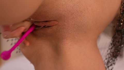 Video of a close-up of a woman's shaved vulva, being groomed with a pink grooming tool. Skin tone is light brown. Background is blurred.