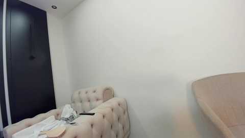 Media: A video of a minimalist, modern bedroom with a beige tufted headboard, a black door, and a small beige table on the right side. The room is sparsely furnished with white walls and a white ceiling.