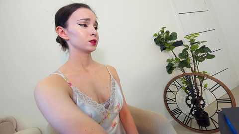 Media: Video of a fair-skinned woman with brown hair, wearing a floral-patterned camisole, sitting on a beige couch against a white wall. Background features a potted plant and a vintage-style clock.