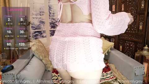 A video of a woman wearing a pink crochet dress, standing in a cozy living room with a plush sofa, wooden screen, and snowy window view.