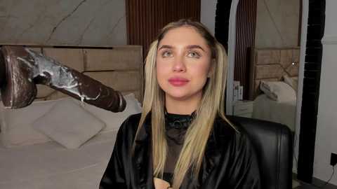 Media: Video of a blonde woman with fair skin, wearing a black salon cape, seated in a modern, upscale salon with marble walls and large mirrors.