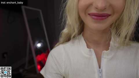 Media: Video of a smiling woman with long blonde hair, wearing a white ribbed zip-up top. Background includes a mirror, red light, and QR code.