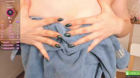 Media: A video of a woman with fair skin, medium build, covering her bare chest with a blue towel. Her green-painted nails are visible. Background includes a TV screen displaying chat messages.