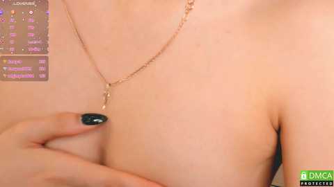 Media: Close-up video of a light-skinned woman with a slim physique, wearing a delicate gold necklace with a small pendant. Her left hand, with dark blue-painted nails, is gently touching her left breast. The background is a soft, neutral tone.