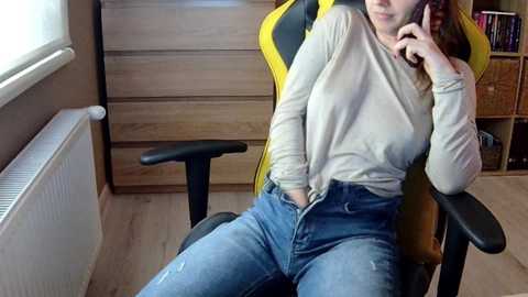 Media: Video of a woman with fair skin, light brown hair, wearing a beige long-sleeve shirt and high-waisted blue jeans, seated in a yellow and black gaming chair, talking on a phone, in a room with wooden furniture and a window.