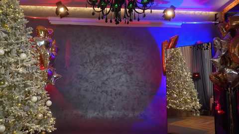 Media: A video of a festive room with two elaborately decorated Christmas trees on either side of a grey, textured wall. The room is lit with warm, blue, and red lights, and a chandelier hangs from the ceiling.